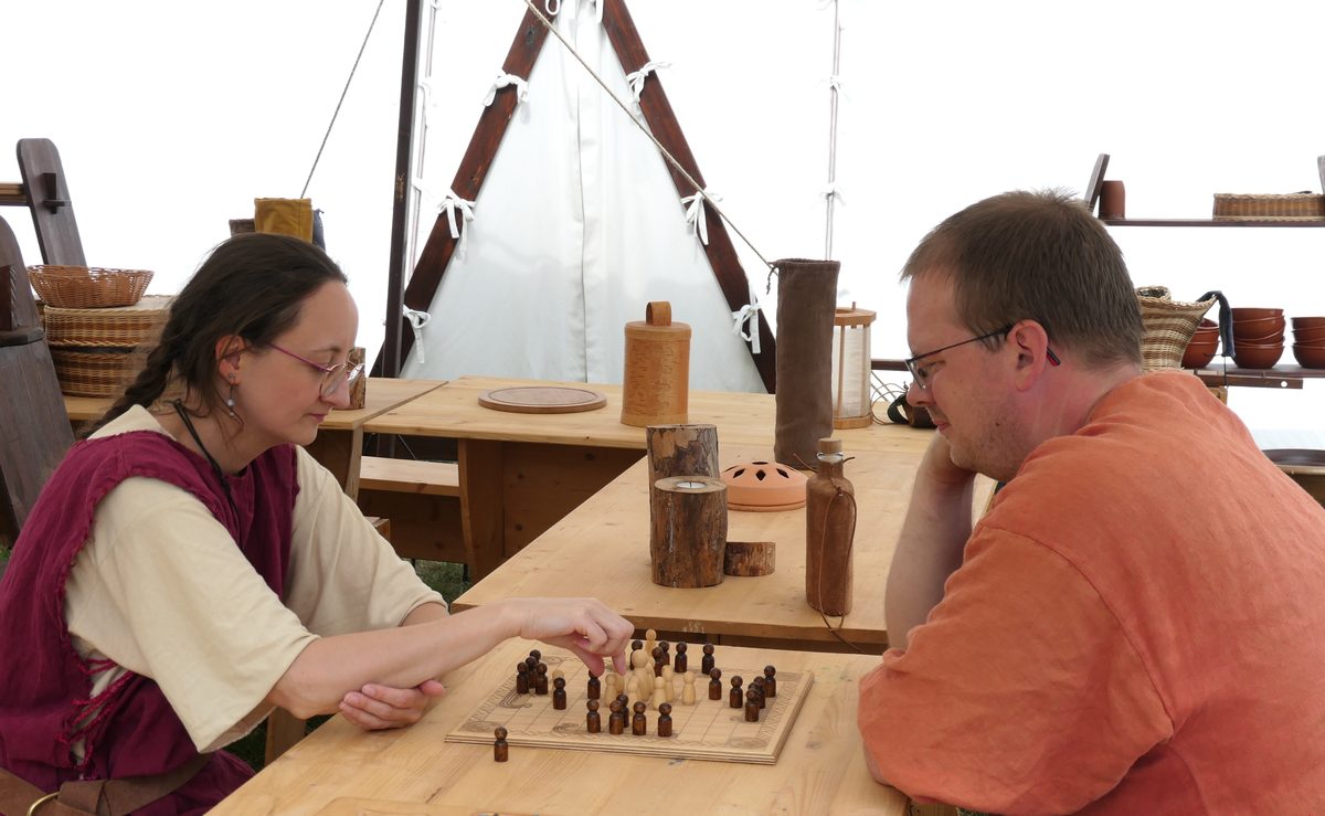 Zwei Gäste in Mittelaltergewandung spielen Hnefatafl an einem Holztisch im Lager von Haus Vingólf; eine Spielerin setzt gerade eine Figur.