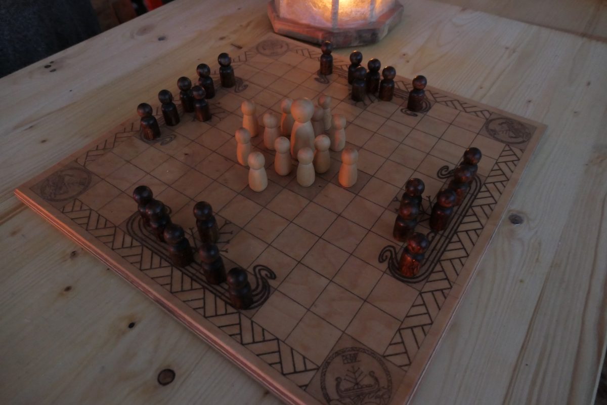 Hnefatafl-Brett in der Abenddämmerung, von einer Oseberg-Lampe beleuchtet; die dunklen und hellen Holzfiguren stehen sich gegenüber.