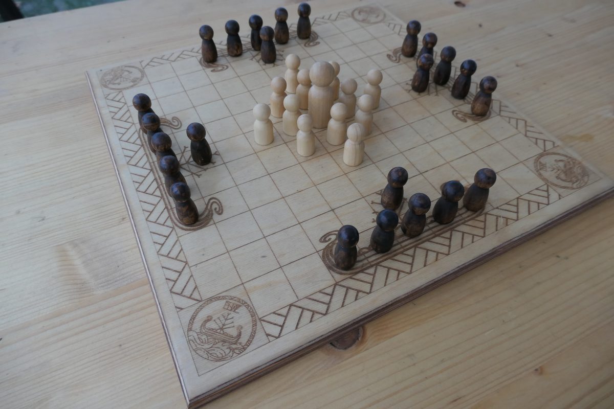 Spielbrett von Vingólf Hnefatafl in Startaufstellung auf einem Holztisch: heller König mit zwölf hellen Verteidigern in der Mitte, 24 dunkle Angreifer in vier Gruppen an den Randmitten.