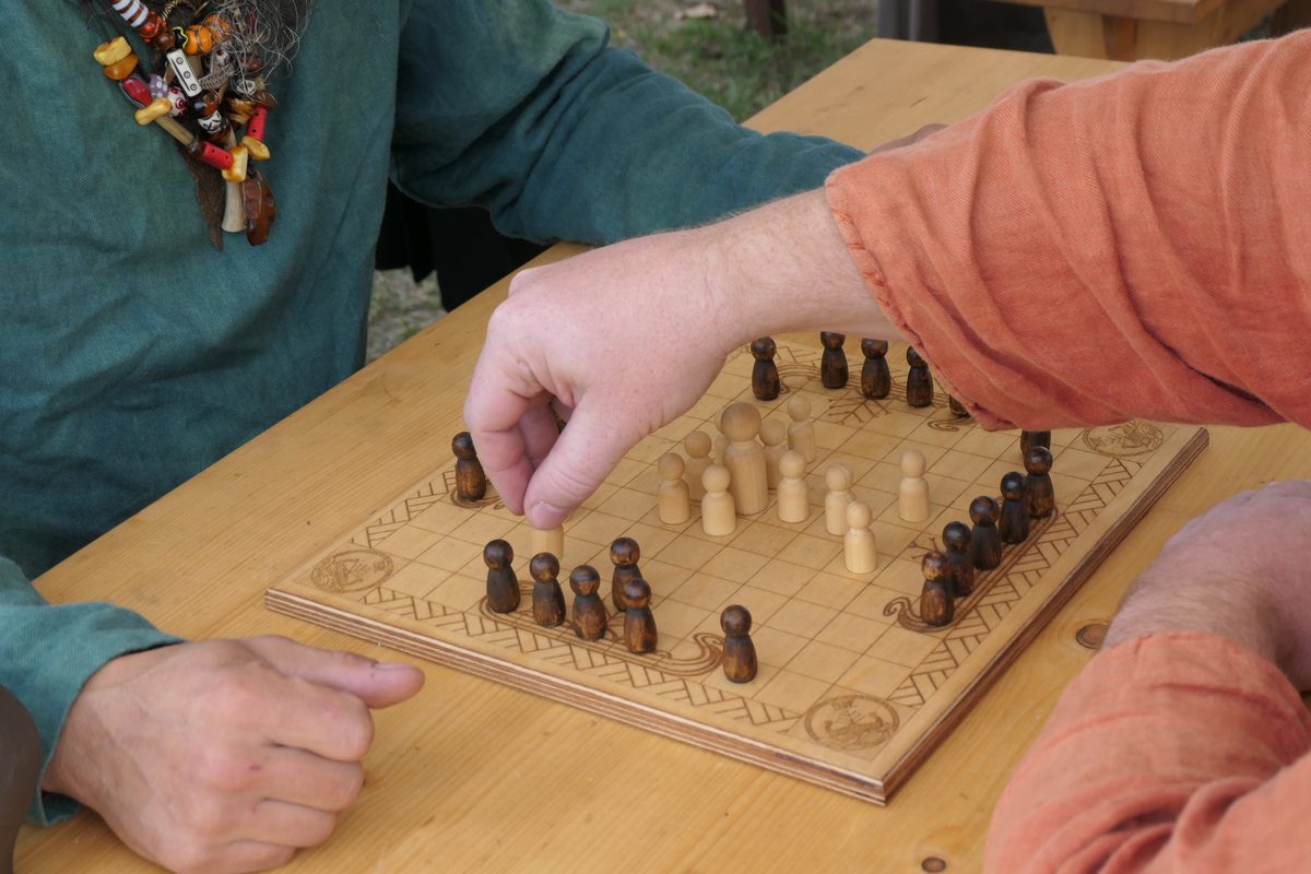 Zwei Spieler in Gewandung sitzen sich an einem Hnefatafl-Brett gegenüber; eine Hand setzt gerade eine helle Figur.
