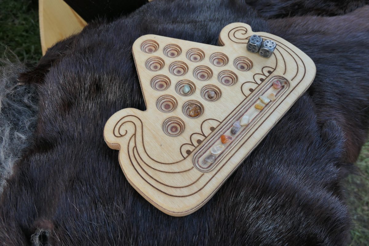 Spielbrett von Kaperfahrt in Form eines Wikinger-Langschiffs aus hellem Holz, mit bunten Glassteinen in den Mulden und zwei Würfeln, auf einem Fell liegend.