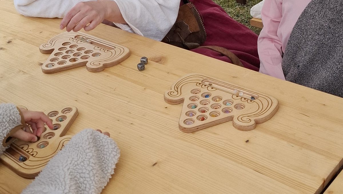 Drei Kaperfahrt-Bretter auf einem Holztisch im Lager. Zwei Hände setzen gerade Glassteine in die Mulden, daneben liegen zwei Würfel.