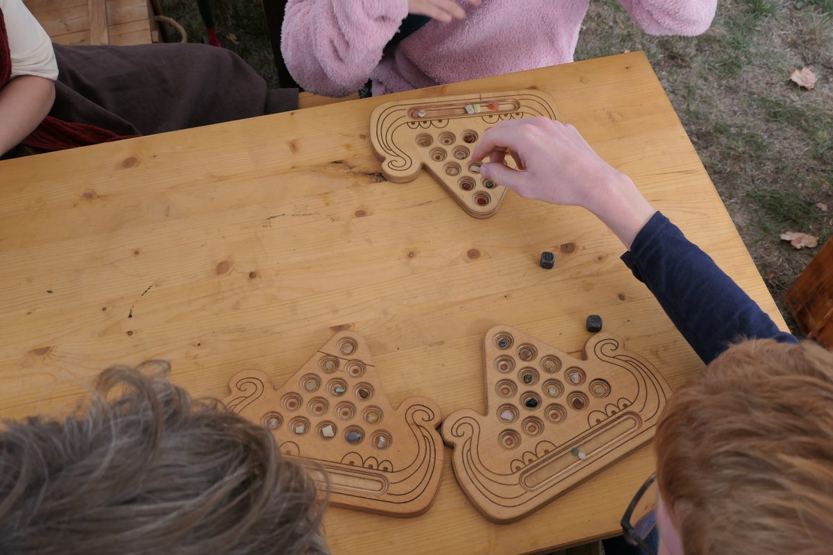 Mehrere Kaperfahrt-Bretter auf einem Holztisch; eine Hand greift über den Tisch hinweg und nimmt einen Stein aus dem Dreieck eines Mitspielers.
