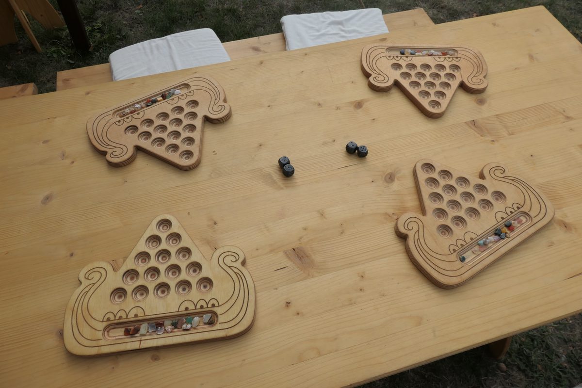 Vier Kaperfahrt-Bretter in Form eines Langschiffs liegen auf einem Holztisch, dazu zwei Würfelpaare; in den Vorratsfeldern liegen bunte Glassteine.