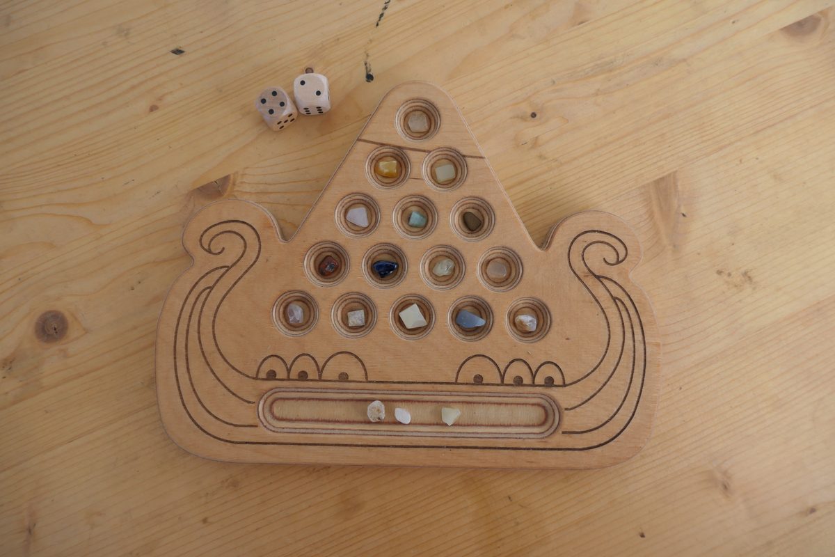 Spielbrett von Kaperfahrt in Form eines Wikinger-Langschiffs aus hellem Holz: ein Dreieck aus 15 runden Mulden, überwiegend mit bunten Glassteinen belegt, darunter im Schiffsbauch das längliche Vorratsfeld, oberhalb zwei Holzwürfel.