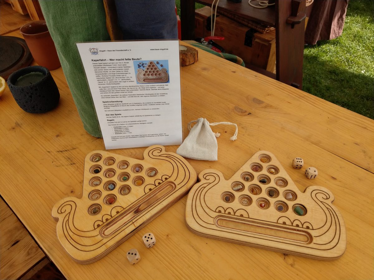 Zwei Kaperfahrt-Bretter auf einem Holztisch im Lager, dazwischen die aufgestellte gedruckte Spielanleitung, ein Leinenbeutel mit Spielsteinen und vier Würfel.